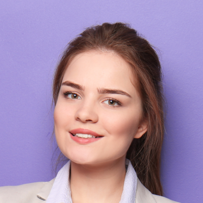 Young woman with brown hair smiling, wearing a light blazer and a striped shirt, posing against a purple background.