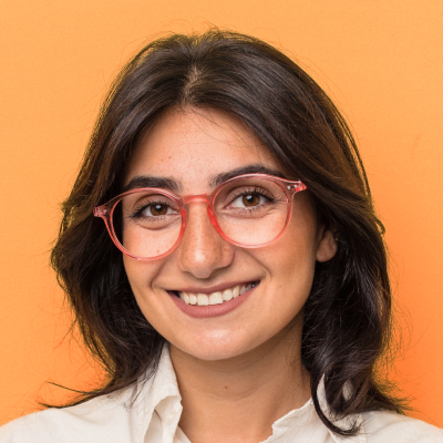 A portrait of a smiling woman with shoulder-length brown hair, wearing red glasses and a white shirt against an orange background.