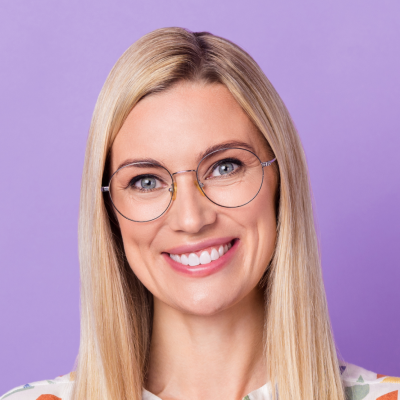 Smiling woman with blonde hair wearing round glasses and a floral shirt, against a purple background.