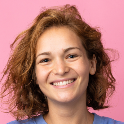 A young woman with curly hair smiling brightly against a pink background. She wears a blue top.