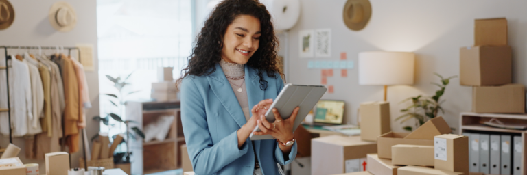 A woman smiles while using a tablet in a creative office space with boxes and fashion items in the background. Biteable video maker